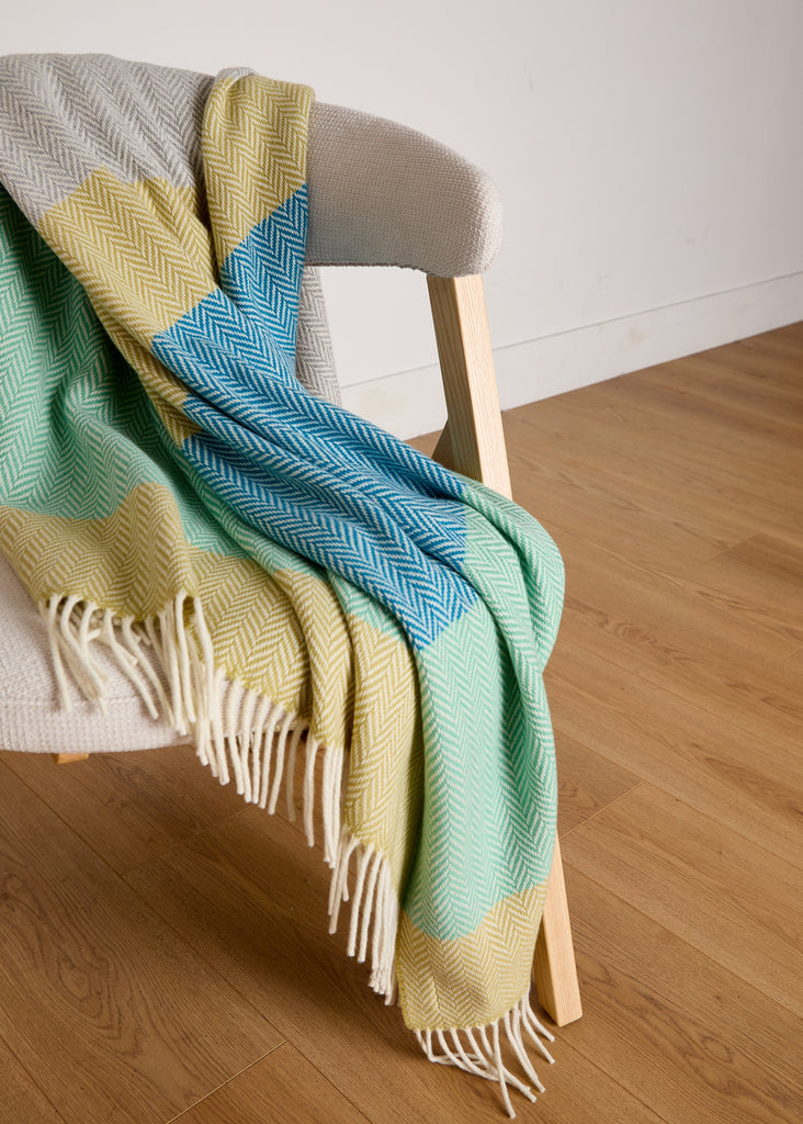 Luxurious Foxford Garavogue Cashmere and Lambswool Throw draped over a modern wooden chair. Featuring a vibrant herringbone pattern in blues, greens, and yellows, with soft fringed edges. Woven in Foxford’s historic mill in County Mayo, Ireland.